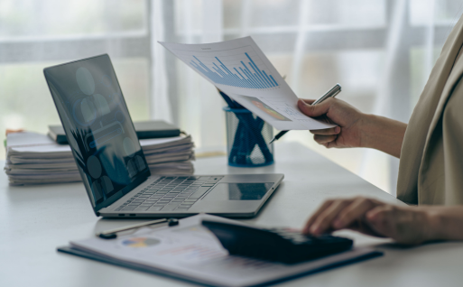 A person is working at a desk, looking at a report with diagrams and holding a pen. In front of them is a laptop with a virtual meeting on the screen. In the foreground is another report with figures and charts.